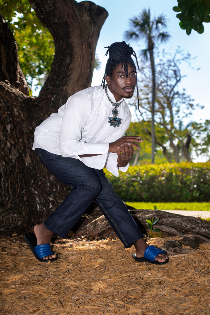 Day Rates for Photographers. Portrait of a Millennial black man with long dreadlocks posing by himself outside standing under a very old oak tree wearing blue jeans, white long sleeve shirt, blue sandals and jewelry.