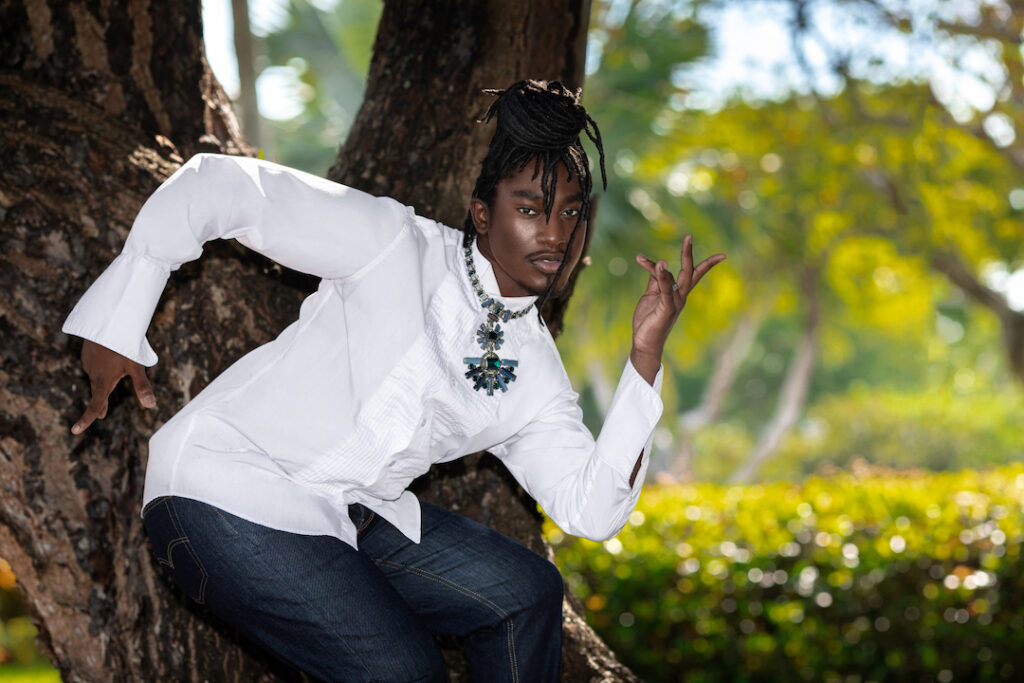 Closeup portrait of a Millennial black man with long dreadlocks posing by himself outside on a summer day dancing under a very old oak tree wearing blue jeans, white long sleeve shirt and jewelry.
