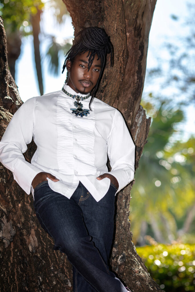 Closeup portrait of a Millennial black man with long dreadlocks posing by himself outside on a summer day leaning on a very old oak tree wearing blue jeans, white long sleeve shirt and jewelry.