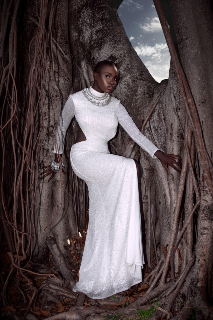 Portrait of a sensual Millennial black woman with bald head and beautiful makeup posing by herself outside barefoot leaning on a very old oak tree wearing a long white dress and gorgeous jewelry.