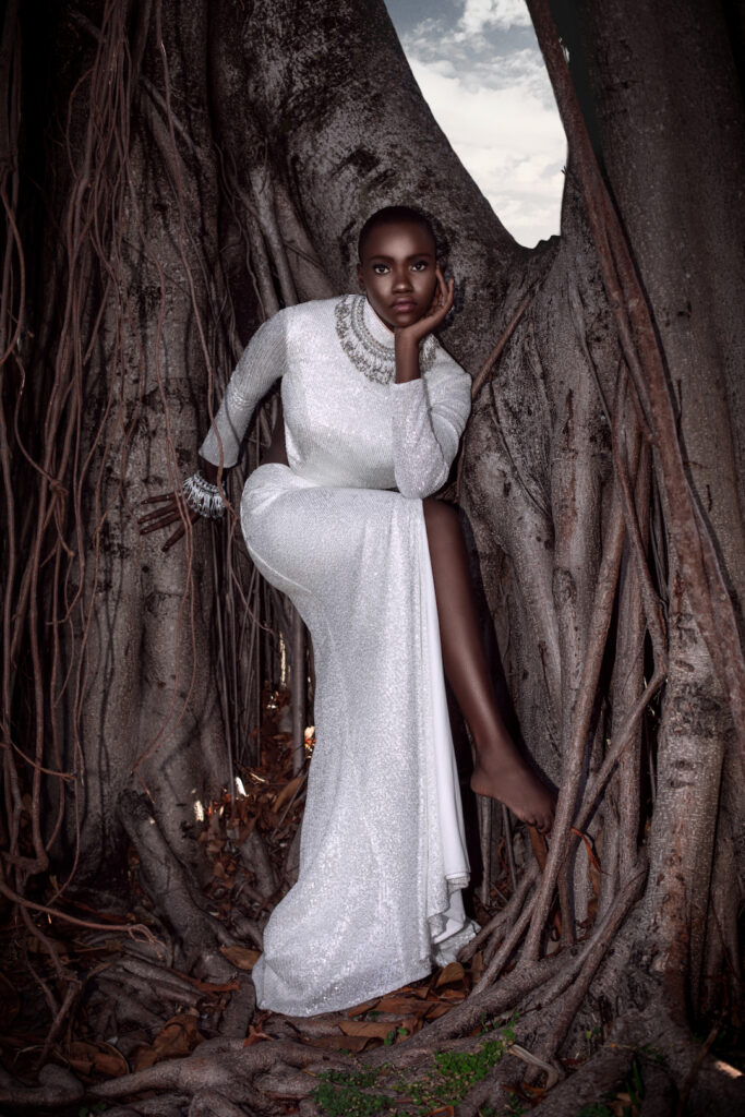 Portrait of a sexy Millennial black woman with bald head and beautiful makeup posing by herself outside barefoot leaning on a very old oak tree wearing a long white dress and gorgeous jewelry.