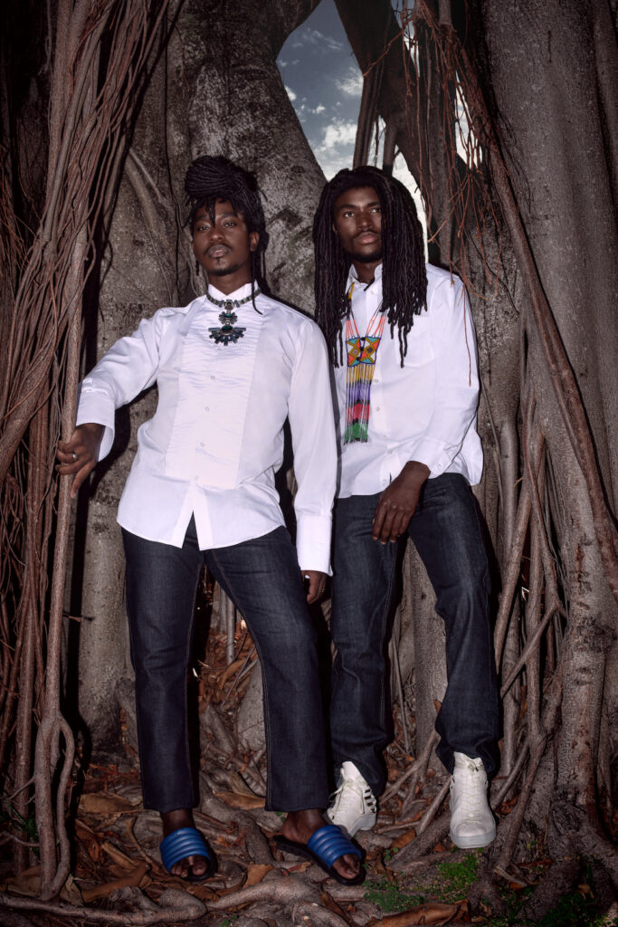 Portrait of two Millennial black man with dreadlocks posing by themselves outside leaning on an old oak tree wearing jeans, long sleeve shirts, gorgeous jewelry with blue sandals and white sneakers.