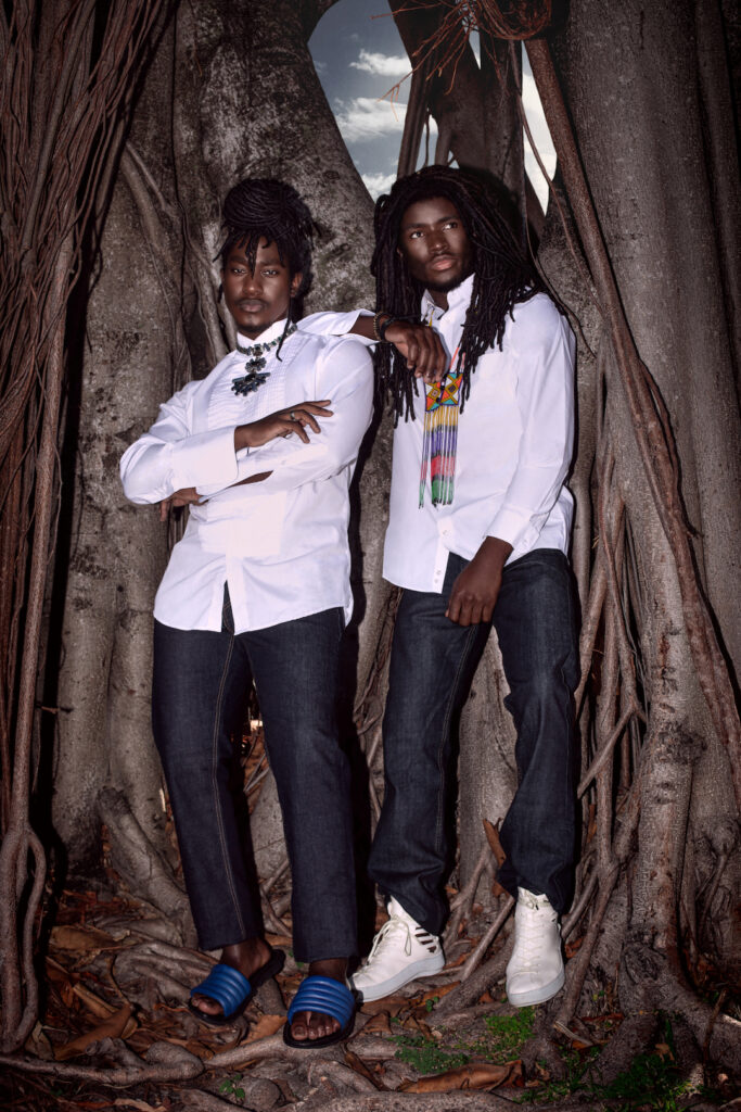 Portrait of two confident Millennial black man with dreadlocks posing by themselves outside leaning on an old oak tree wearing jeans, long sleeve shirts, jewelry with blue sandals and white sneakers.