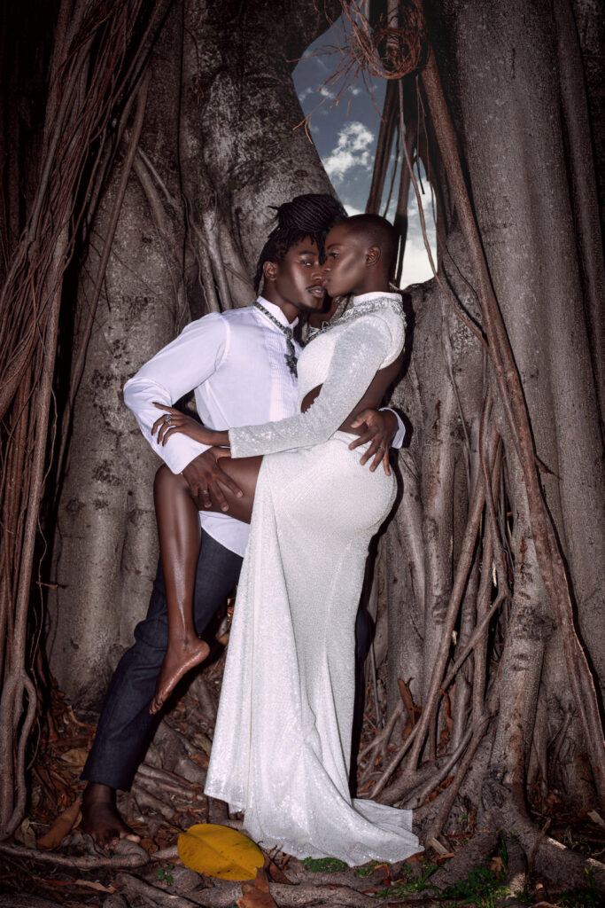Portrait of a sensual Millennial black couple posing by themselves outside barefoot leaning on an old oak tree on a summer day wearing jeans, long sleeve shirt and a long white dress with jewelry.