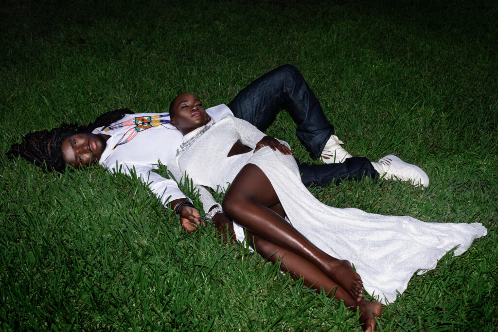 Portrait of a sensual Millennial black couple posing by themselves outside laying on grass on a summer evening wearing jeans, long sleeve shirt with sneakers and a long white dress with jewelry.