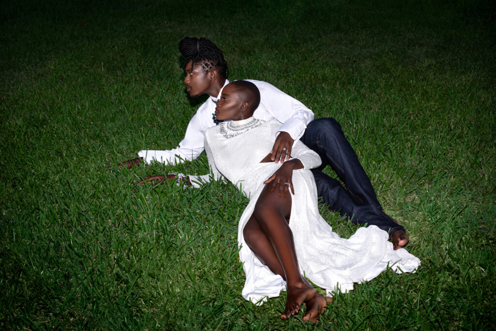 Portrait of a serene Millennial black couple posing by themselves outside laying on grass on a summer evening wearing jeans, long sleeve shirt with sneakers and a long white dress with jewelry.