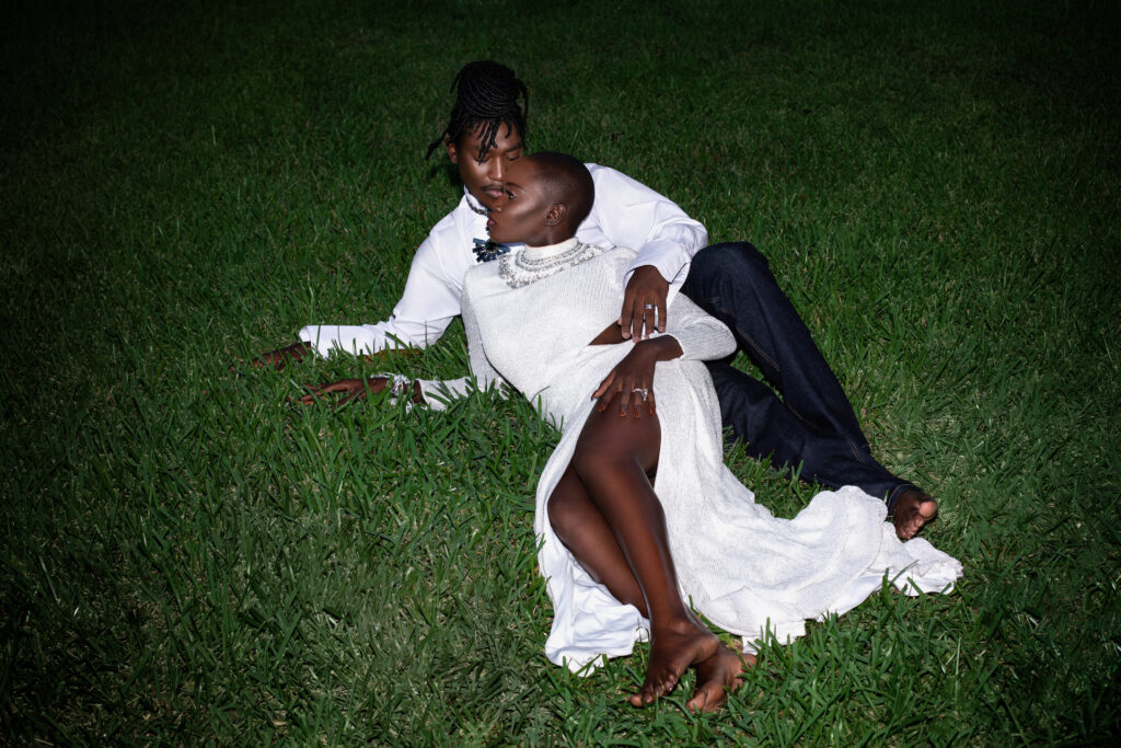 Portrait of an intimate Millennial black couple posing by themselves outside laying on grass barefoot on a summer evening wearing jeans, long sleeve shirt and a long white dress with jewelry.