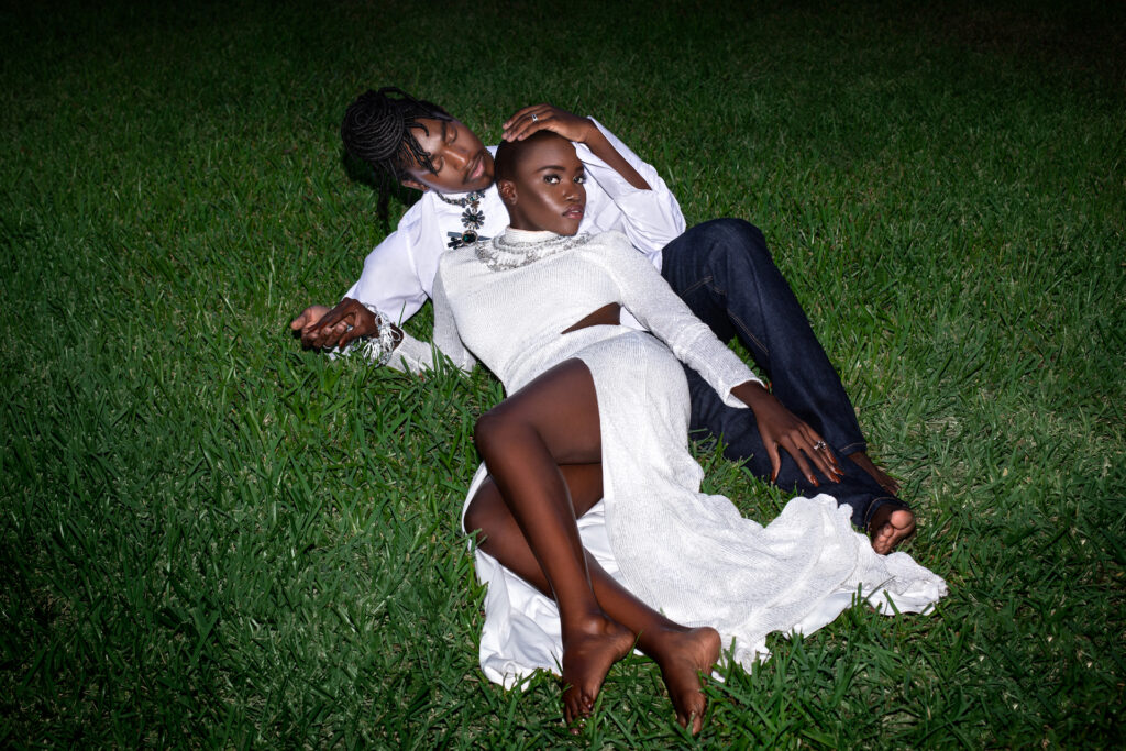 Portrait of a sexy Millennial black couple posing by themselves outside laying on grass barefoot on a summer evening wearing jeans, long sleeve shirt and a long white dress with jewelry.