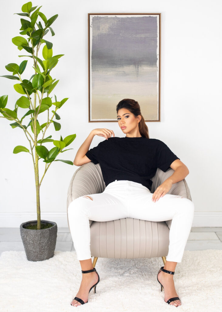 Become your own best friend. Portrait of a confident young Latina with long wavy hair and beautiful makeup sitting in a grey leather lounge chair inside a living room with carpet wearing white pants, a black top and high heels.