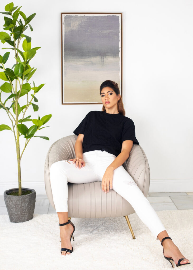 Portrait of a sensual young Latina with long wavy hair and beautiful makeup sitting in a grey leather lounge chair inside a living room with carpet wearing white pants, a black top and high heels.