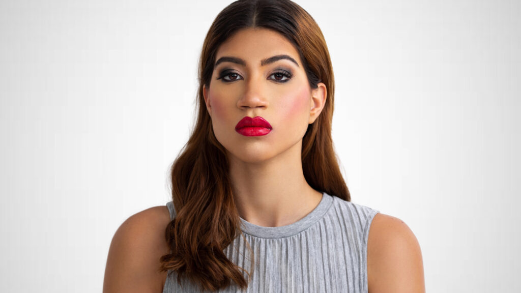 Closeup portrait of a sensual young Latina with long wavy hair over her left shoulder, perfect eye brows, dark eye makeup and red lipstick wearing a grey bare shoulder dress.