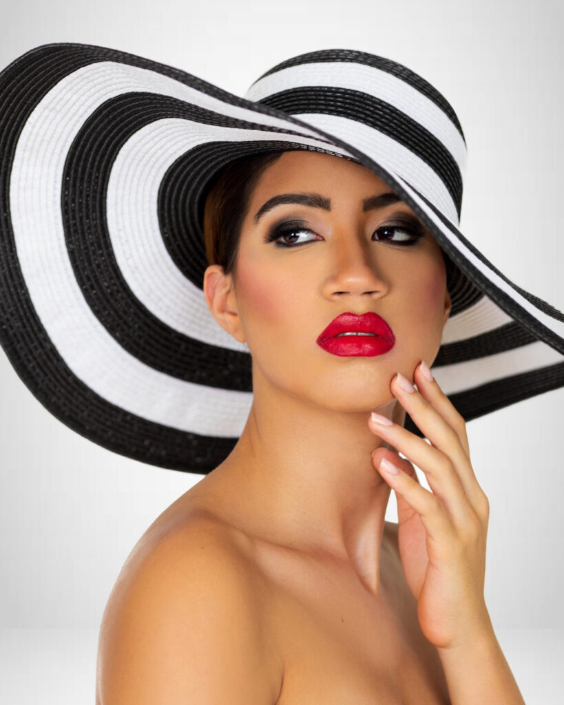 Closeup portrait of a serene young Latina with long wavy hair in an up-do, perfect eye brows, dark eye makeup and red lipstick wearing a large black and white striped summer hat.