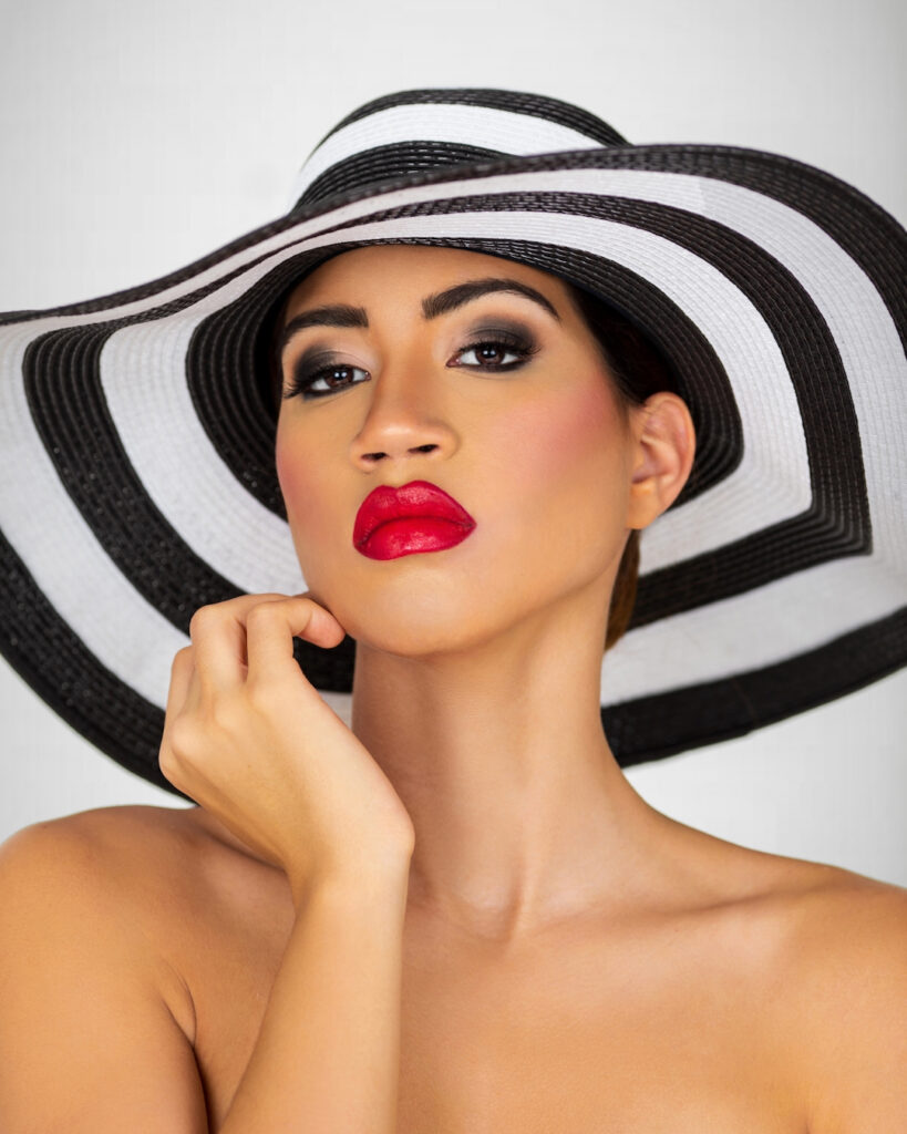 Become your own best friend. Closeup portrait of a sensual young Latina with long wavy hair in an up-do, perfect eye brows, dark eye makeup and red lipstick wearing a large black and white striped summer hat.
