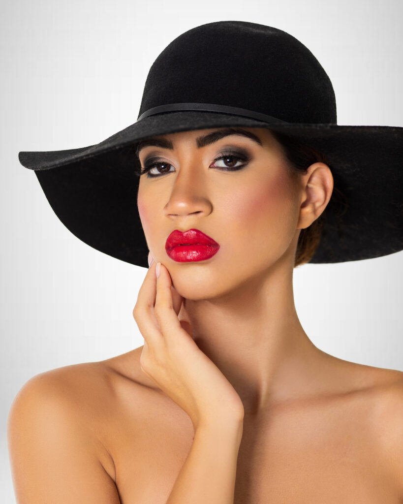 Closeup portrait of a sensual young Latina with long wavy hair in an up-do, perfect eye brows, dark eye makeup and red lipstick wearing a large black hat.