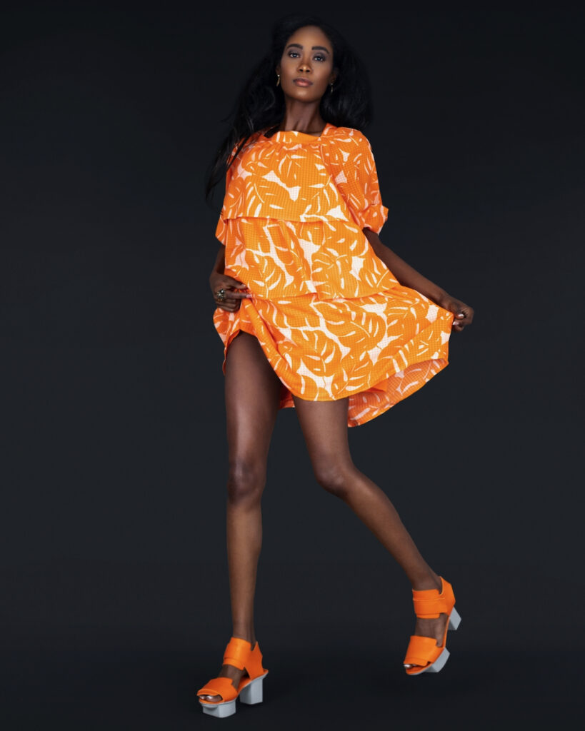 Boho confidence. Portrait of a sensual mature black woman with long hair, beautiful makeup posing by herself in a studio with a dark background wearing a short flowy orange dress with orange shoes and jewelry.