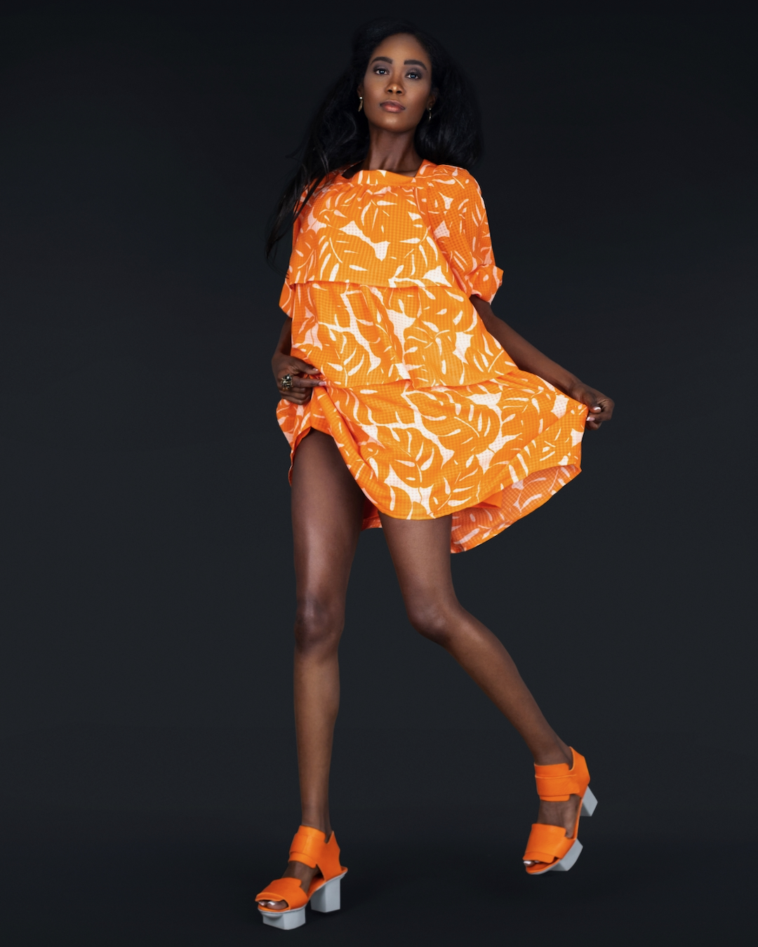 Maria wearing a short flowing orange dress with matching platform shoes and statement jewelry