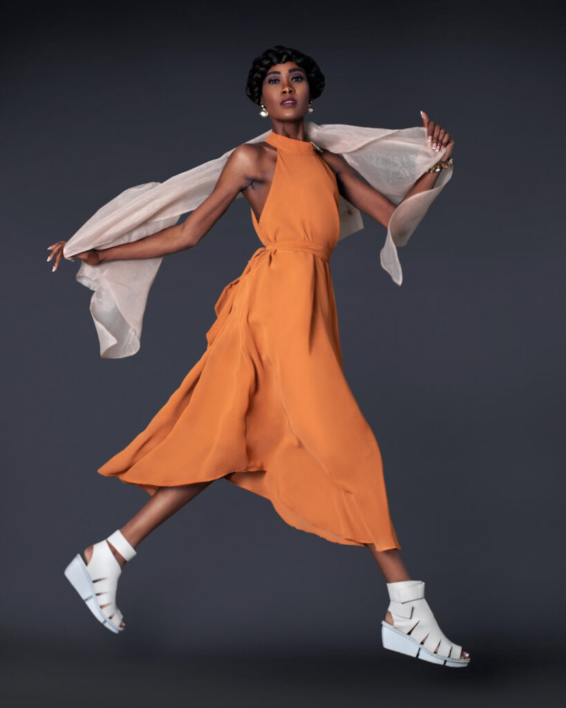 Portrait of a sensual black woman with long hair, beautiful makeup floating in midair by herself in a studio with a grey background wearing a long flowy orange dress with white shoes and jewelry.