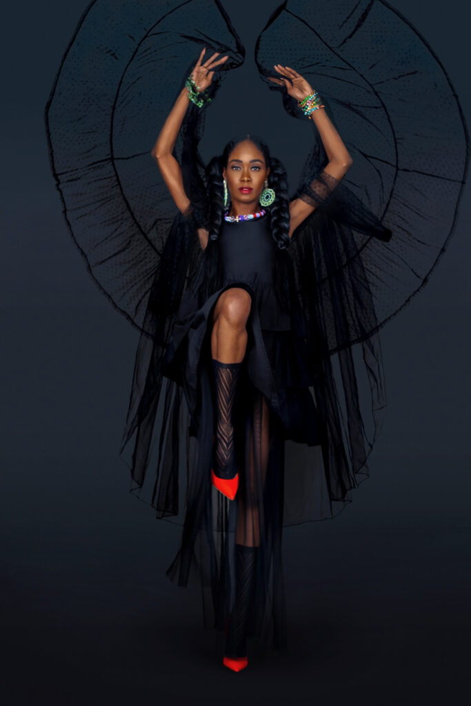 Boho confidence. Portrait of a beautiful black woman with long hair, beautiful makeup posing by herself in a studio with a dark background wearing a long flowy black dress with red high heels and jewelry.