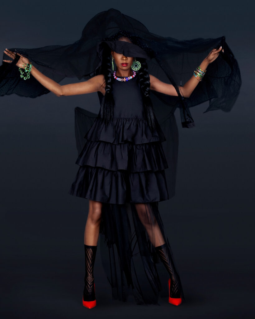 Portrait of a serene black woman with long hair, beautiful makeup posing by herself in a studio with a dark background wearing a long flowy black dress with red high heels and jewelry.