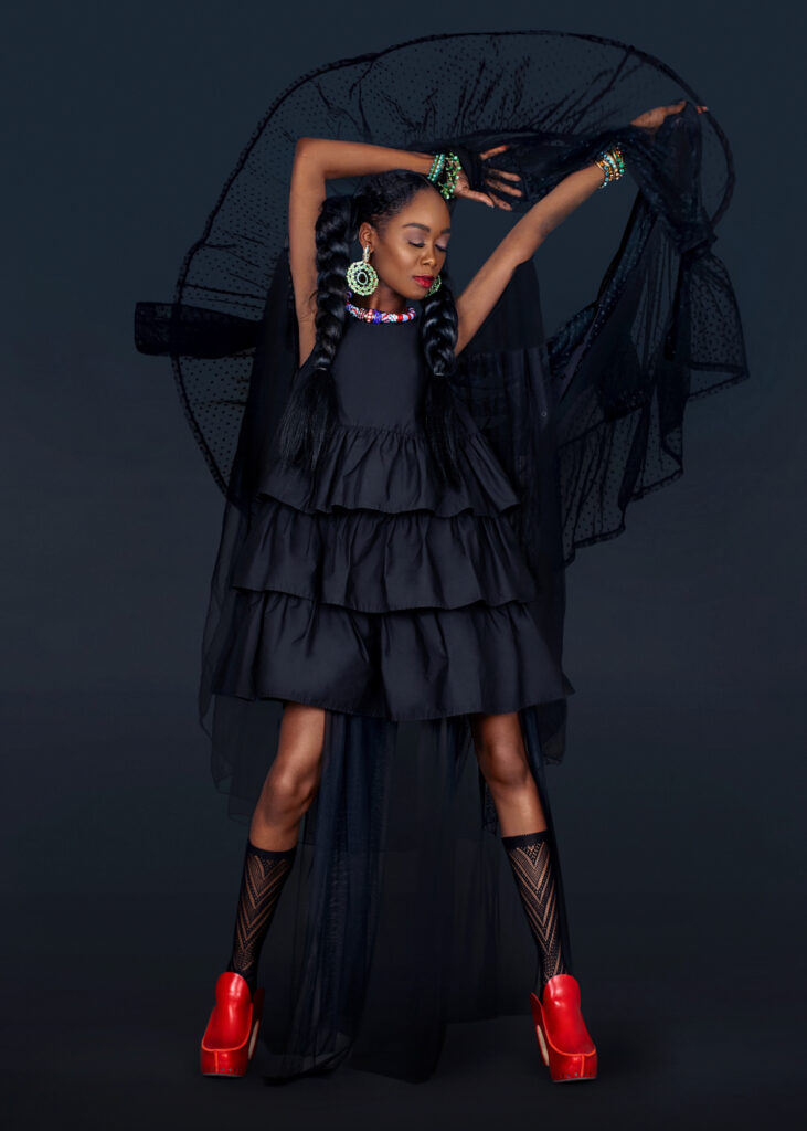 Portrait of a sensual black woman with long hair, beautiful makeup posing by herself in a studio with a dark background wearing a long flowy black dress with red shoes and jewelry.