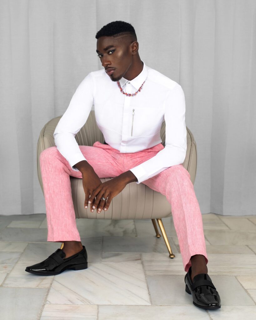 Portrait of a Black man in pink pants and white shirt sitting in a grey leather lounge chair — Pretty Boy Joy editorial.