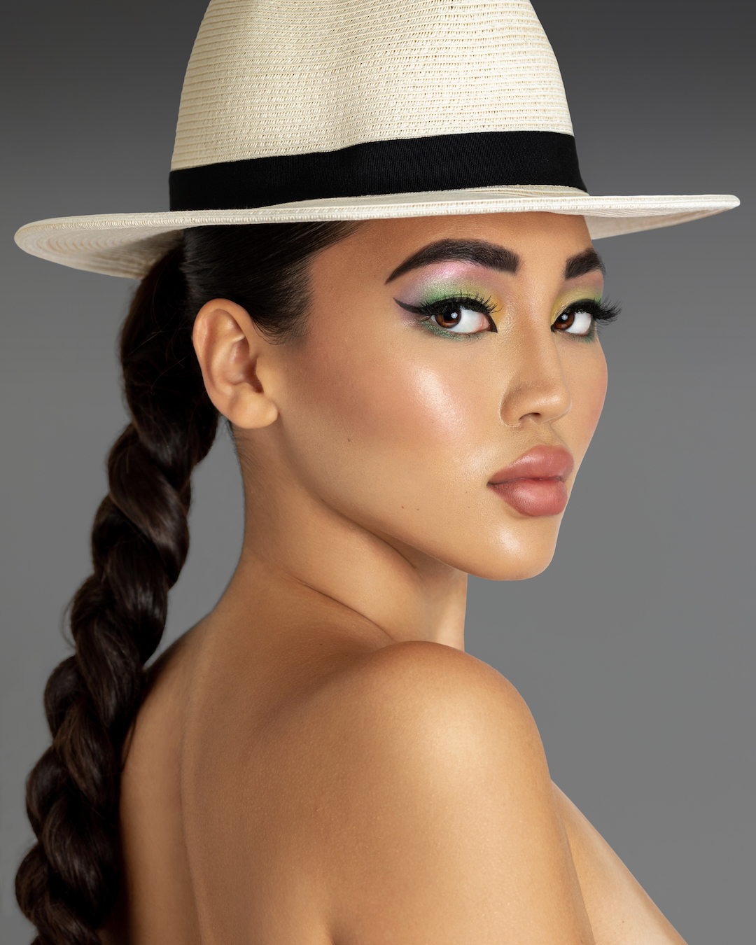 Crazy sexy confidence. Closeup portrait of a sensual young Asian woman with long braided ponytail hair and beautiful makeup standing by herself inside a studio with a grey background wearing a white fedora hat.