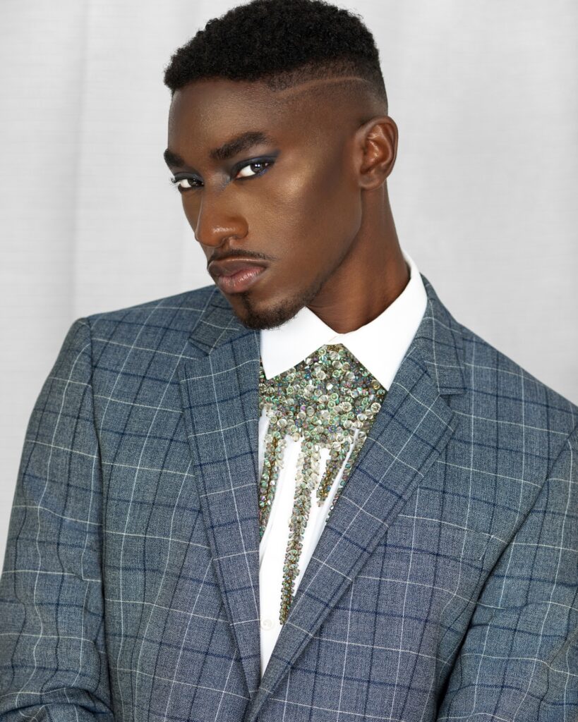 Closeup portrait of a sensual Black man with faded Afro hair and beautiful makeup posing by himself inside a living room with curtain background wearing blue suit with a white shirt and jewelry.