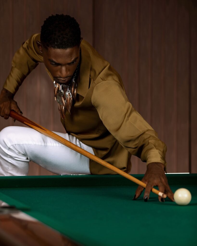 Portrait of a confident Black man with short Afro hair posing by himself inside a pool billiards hall wearing a brown button up shirt with white denim jeans and a long necklace.