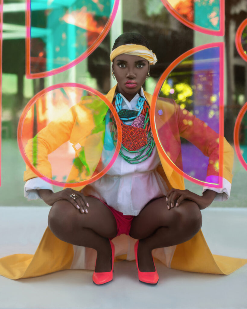 An elegant young black woman with short black hair & beautiful makeup and gorgeous jewelry posing by herself outdoors kneeling inside a glass box is wearing a yellow coat, white blouse & red shorts.