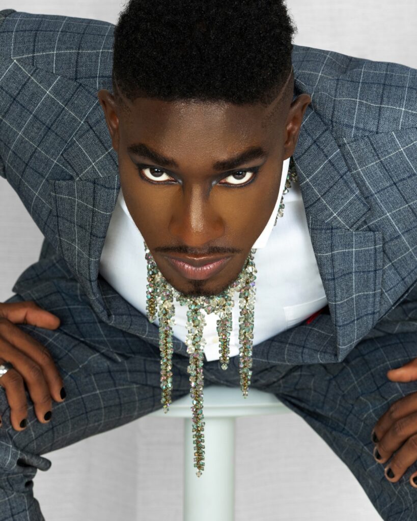 Portrait of a handsome Black man with faded Afro hair and beautiful makeup sitting on a grey bar chair inside a living room with curtain background wearing blue suit, white shirt and jewelry.