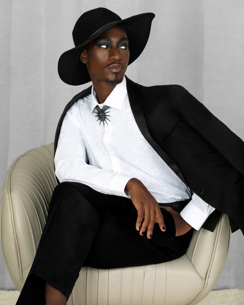 Serene Black man wearing a black suit, hat and jewelry on a grey lounge chair — Pretty Boy Joy editorial.