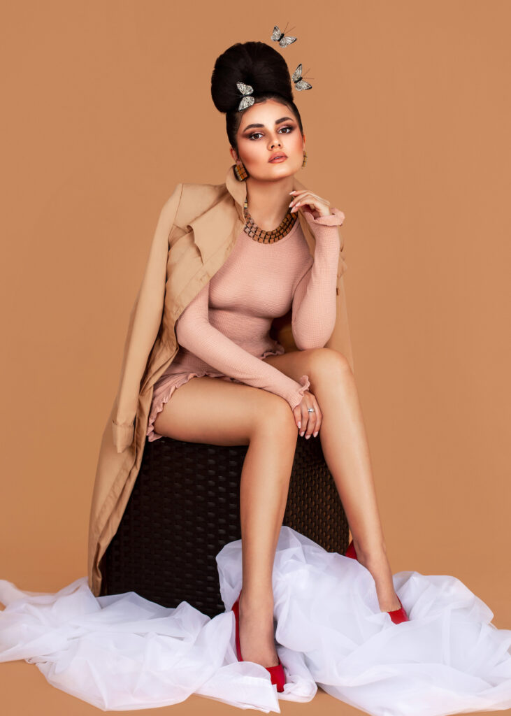 Portrait of a sexy Latin girl with long wavy hair in an up-do and beautiful makeup sitting by herself surrounded by butterflies inside a studio with pecan color background wearing a short pink outfit.