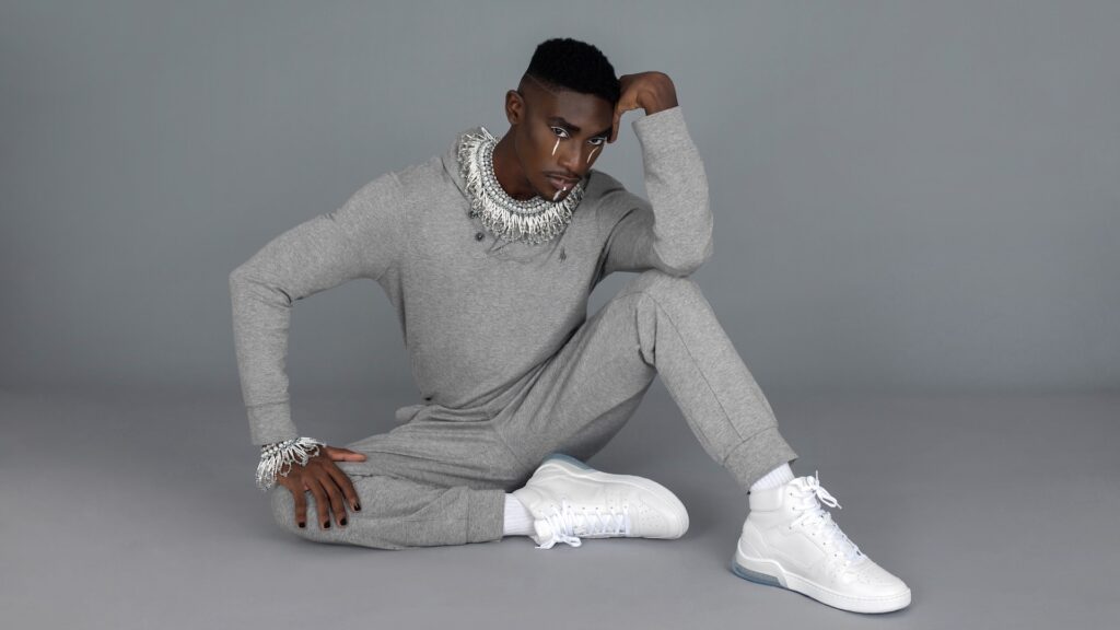 Sensual Black man in a grey tracksuit and white sneakers sitting on the studio floor — Pretty Boy Joy editorial.
