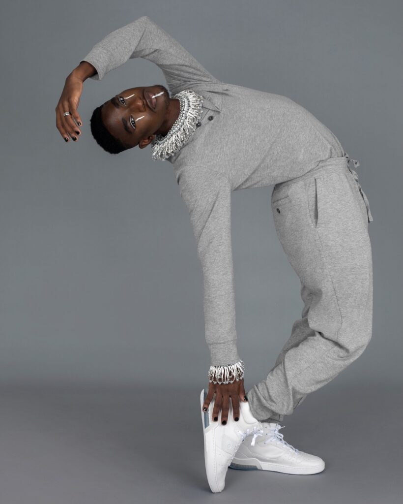 Portrait of a confident Black man with faded Afro hair and beautiful makeup posing by himself inside a studio with grey background wearing a grey track suit, white sneakers and jewelry.