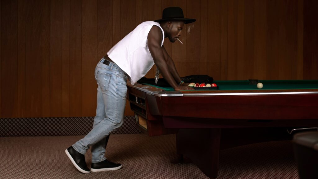 Pool Rocker Show Stopper. Portrait of a masculine Black man with a fedora hat posing by himself inside a pool billiards hall wearing a black leather jacket, white T-shirt with with blue denim jeans and a long necklace.