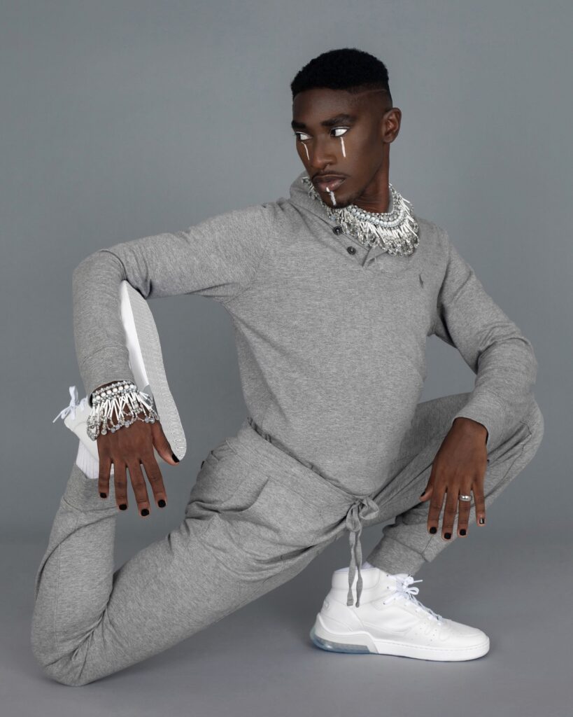 Portrait of a serene Black man with faded Afro hair and beautiful makeup posing by himself inside a studio with grey background wearing a grey track suit, white sneakers and jewelry.