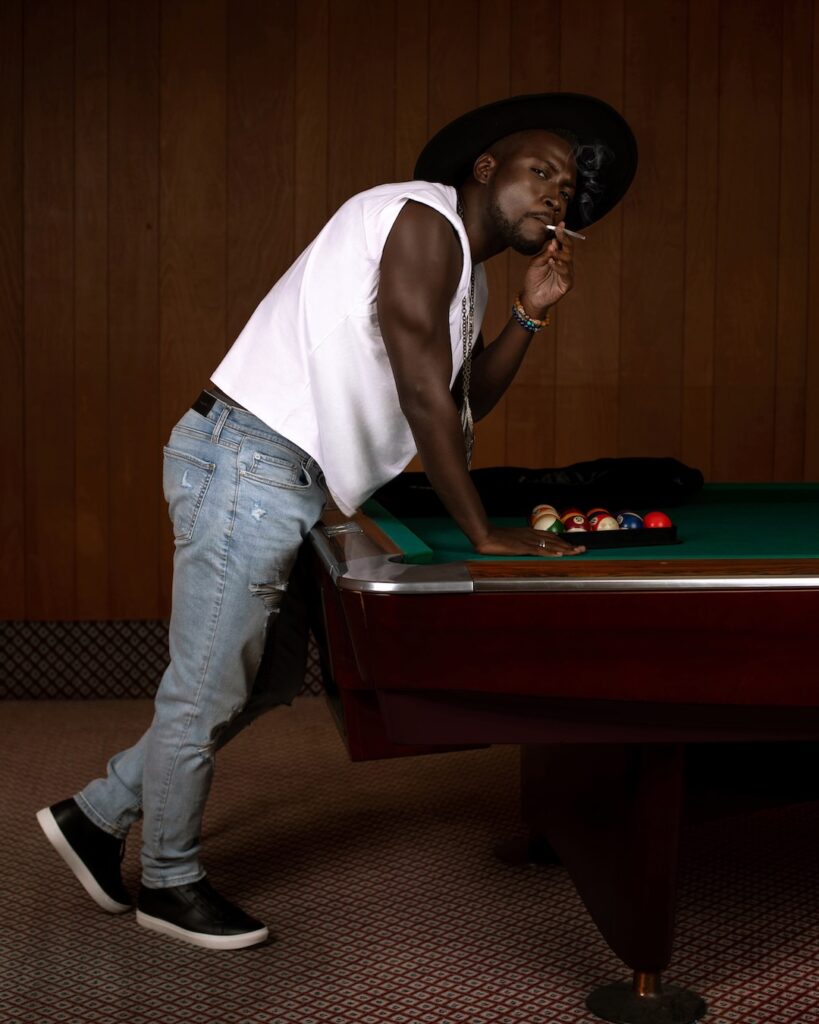 Portrait of a cool Black man with a fedora hat posing by himself inside a pool billiards hall wearing a black leather jacket, white T-shirt with blue denim jeans and a long necklace.