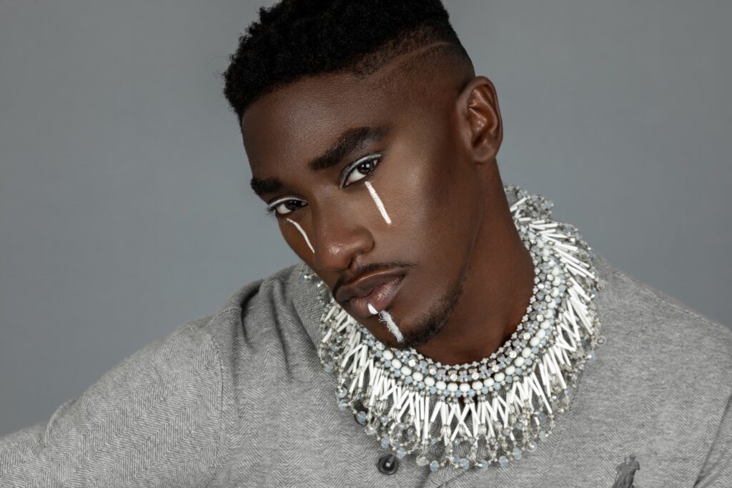 Closeup portrait of a handsome Black man with faded Afro hair and beautiful makeup posing by himself inside a studio with grey background wearing a grey track suit and jewelry.