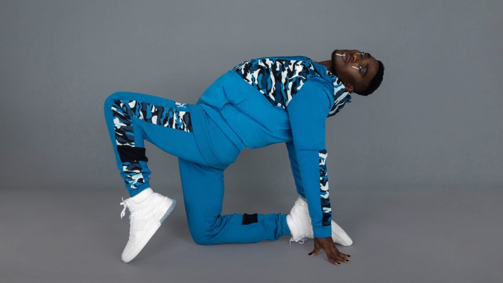 Confident Black man posing in a blue tracksuit and white sneakers against a grey studio backdrop — Pretty Boy Joy editorial.