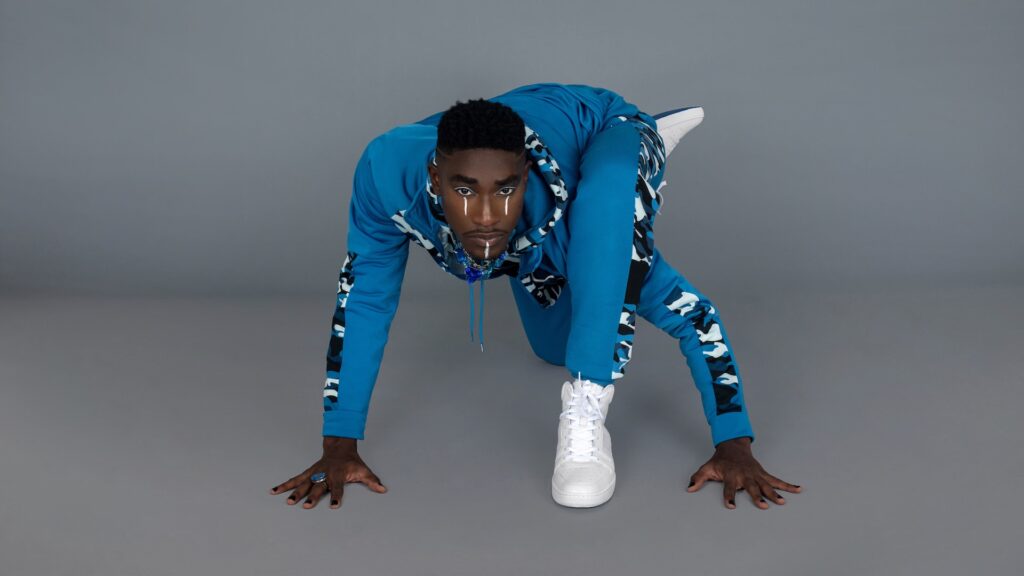 Portrait of a strong Black man with faded Afro hair and beautiful makeup posing by himself inside a studio with grey background wearing a blue track suit, white sneakers and jewelry.