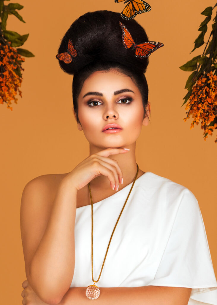 Portrait of a strong Latin girl with long wavy hair in an up-do and beautiful makeup posing by herself surrounded by flowers and butterflies inside a studio with pecan color background.