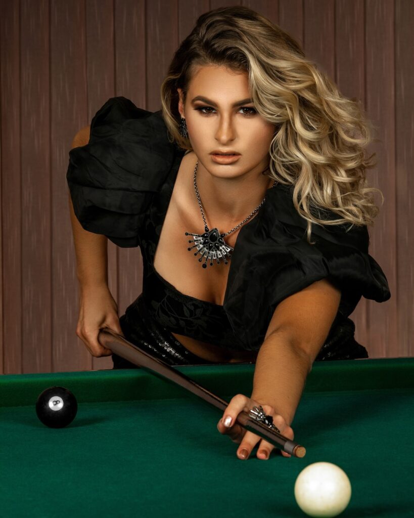 Closeup portrait of a focused White woman with long curly blonde hair and beautiful makeup posing by herself playing pool billiards wearing a short black top with a black diamond necklace and ring.