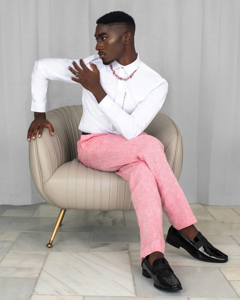 Portrait of a serene Black man with faded Afro hair and beautiful makeup sitting in a grey leather lounge chair inside a living room wearing pink pants, a white shirt and black leather shoes.