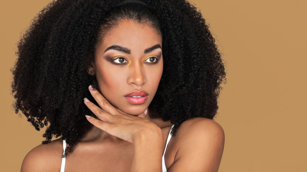 Modern beauty and self-expression. Katherine with long dark hair, eye makeup, white strap top, studio with pecan background.