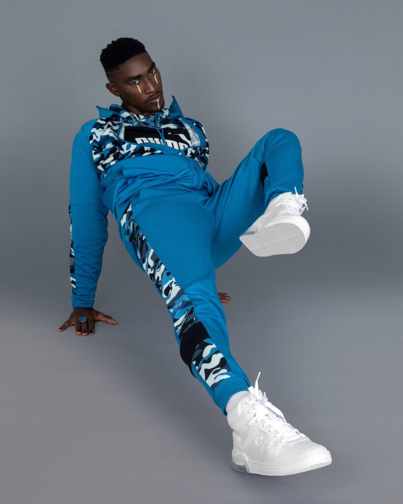 Portrait of a sensual Black man with faded Afro hair and beautiful makeup posing by himself inside a studio with grey background wearing a blue track suit, white sneakers and jewelry.