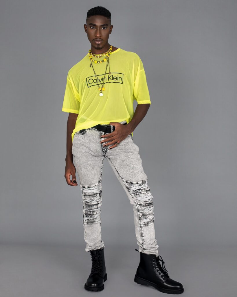 Portrait of a handsome Black man with faded Afro hair and beautiful makeup standing by himself inside a studio with grey background wearing a yellow mesh top, grey jeans, black boots and jewelry.