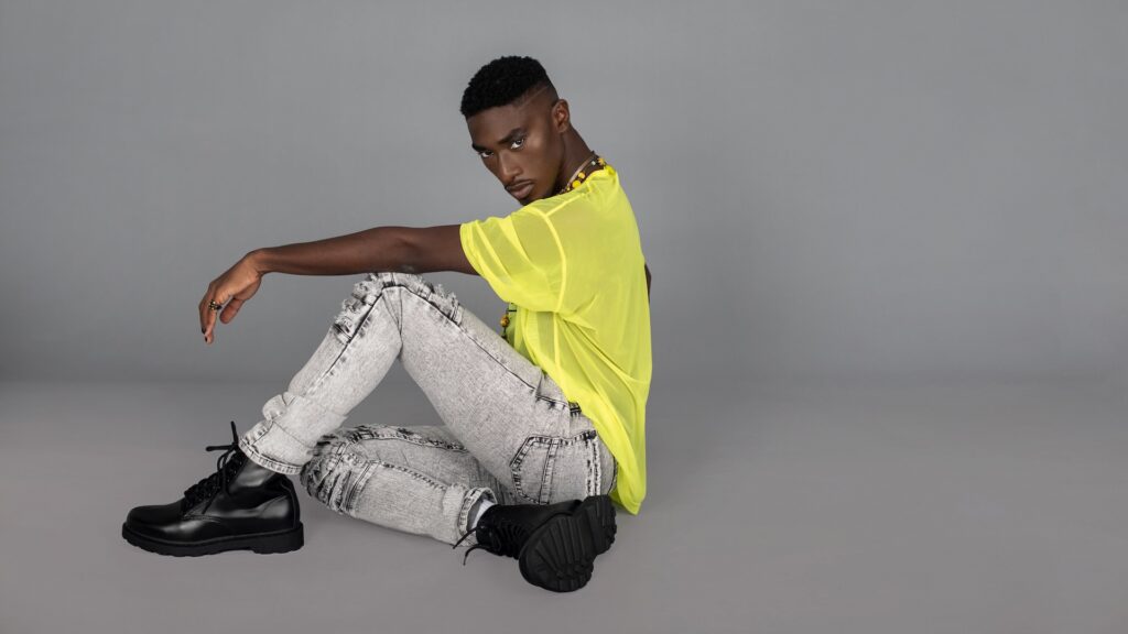 Portrait of a sensual Black man with faded Afro hair and beautiful makeup sitting by himself inside a studio with grey background wearing a yellow mesh top, grey jeans, black boots and jewelry.