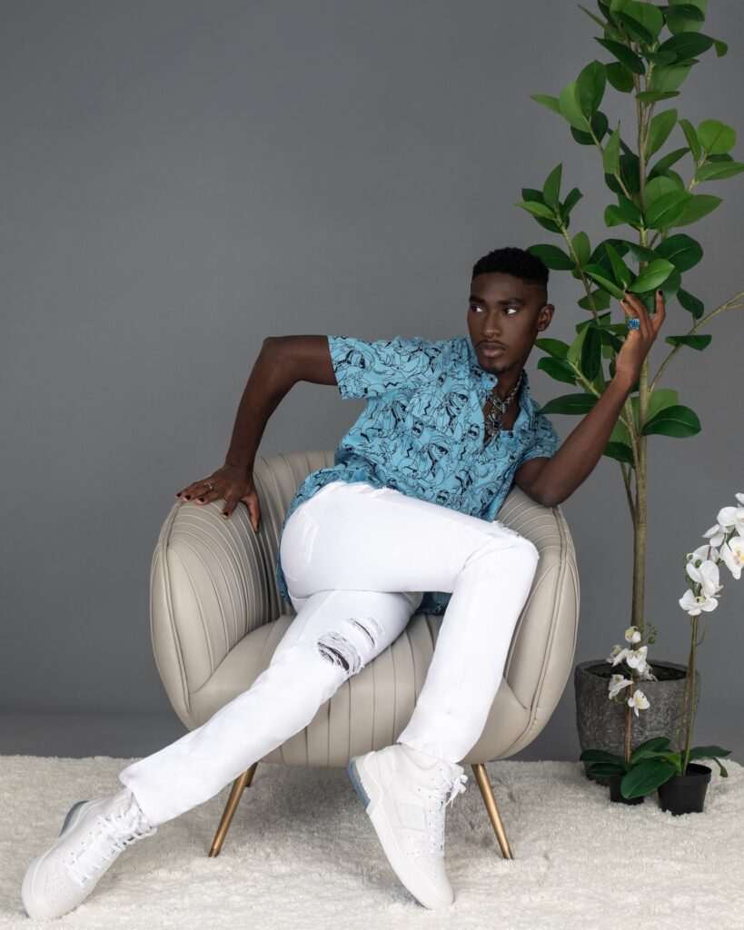 Portrait of a serene Black man with faded Afro hair and beautiful makeup sitting by himself in a grey lounge chair indoors wearing a blue button shirt, white jeans, white sneakers and jewelry.