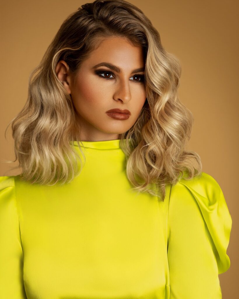 Closeup portrait of a serene White woman with long curly blonde hair and beautiful makeup posing by herself inside a studio with beige background wearing a yellow top.