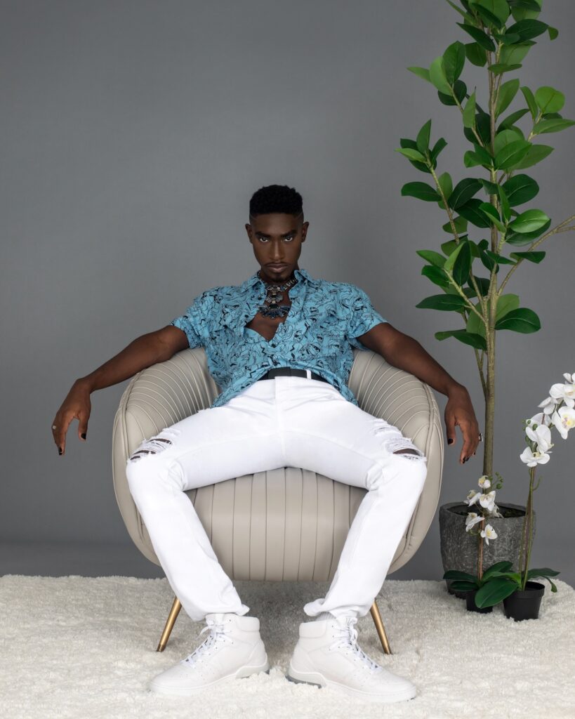 Portrait of a confident Black man with faded Afro hair and beautiful makeup sitting by himself in a grey lounge chair indoors wearing a blue button shirt, white jeans, white sneakers and jewelry.